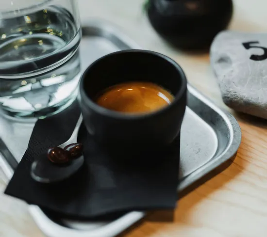 Espresso in schwarzer Tasse mit Wasserglas auf Metalltablett und schokolierten Bohnen